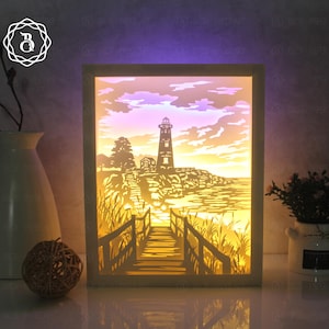 Puede incluir: Un marco de madera blanco con un diseño de papel recortado iluminado de un faro en una costa rocosa con un puente de madera que conduce a él. La escena está iluminada con luz blanca cálida.