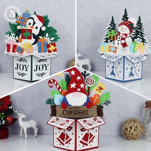 May include: Three Christmas-themed pop-up box cards. One features a penguin with gifts, another a snowman, and the third a gnome. Each card has a box base with festive designs and the words "JOY" or "MERRY CHRISTMAS".