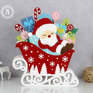 May include: A festive Christmas decoration featuring a sleeping Santa Claus in a red sleigh. The sleigh is adorned with white snowflakes and trees. The sleigh is filled with wrapped gifts, candy canes, and lollipops. The Santa Claus figure is wearing a red suit and hat with a white beard.
