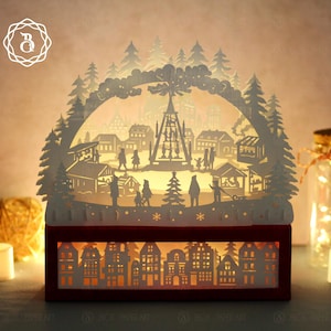 Puede incluir: Una escena decorativa de un pueblo navideño, elaborada en papel blanco, se ilumina desde el interior de una base de madera roja. El intrincado diseño presenta siluetas de casas, árboles y personas, creando un ambiente festivo. La escena está iluminada por una luz cálida.