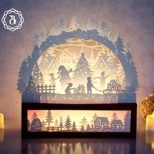 Peut inclure: Une scène de Noël en papier blanc représentant le Père Noël, des enfants et un traîneau. La scène est éclairée de l'intérieur, montrant un village d'hiver avec des maisons et des arbres. La base est un rectangle marron foncé.