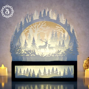 Può includere: Un'opera d'arte in carta a strati raffigurante una scena di foresta con cervi e alberi, illuminata dall'interno. L'opera presenta una base nera e una silhouette di foresta bianca, creando un bagliore caldo. Il design include un logo nell'angolo in alto a sinistra.