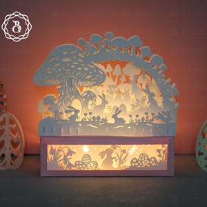 Puede incluir: Una caja de luz con temática de Pascua cortada en papel blanco. El diseño presenta conejos, setas y huevos de Pascua. La caja de luz es rectangular con un marco rosa y un brillo cálido que emana del interior. Se ven huevos de Pascua decorativos.