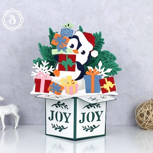 May include: A Christmas-themed pop-up card with a penguin in a Santa hat, surrounded by gifts and green foliage. The box displays the word "JOY" in green. White snowflakes and a white deer figurine are also visible.