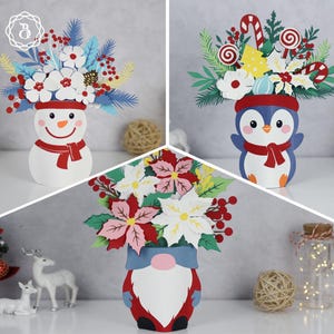 May include: Three festive paper flower arrangements. One features a snowman with a red scarf, another a penguin with a red scarf, and the last a gnome with a blue hat. Each arrangement has colorful paper flowers and holiday accents.
