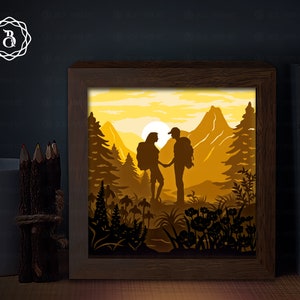 Puede incluir: Una obra de arte de papel recortado en un marco de madera que representa a una pareja haciendo senderismo en un paisaje montañoso. La escena está iluminada desde atrás, creando un brillo cálido. La pareja está silueteada contra la puesta de sol, con montañas y árboles en el fondo.