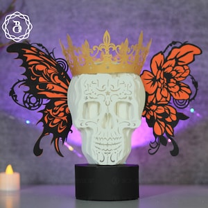Puede incluir: Una escultura de calavera de papel en capas con una corona dorada y alas de mariposa naranjas y negras. La calavera es blanca y las alas están intrincadamente cortadas. La escultura se asienta sobre una base negra, creando una pieza decorativa única.