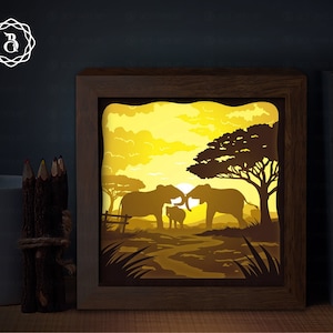 Peut inclure: Une œuvre d'art en découpe de papier représentant une famille d'éléphants dans un décor de savane. Les éléphants sont en silhouette contre un ciel jaune de coucher de soleil avec un grand arbre en arrière-plan. L'œuvre d'art est encadrée dans un cadre en bois brun.