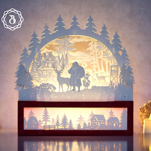Pode incluir: Decoração de Natal iluminada com uma cena de inverno em camadas. Apresenta o Pai Natal, renas e um boneco de neve. A base é um retângulo vermelho escuro, e o design geral é branco.