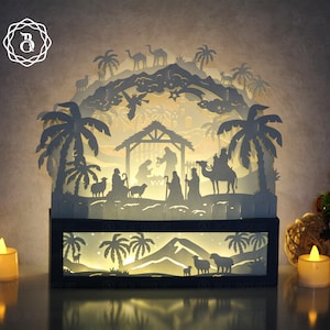 Peut inclure: Une crèche de Noël en papier découpé en couches, avec une base noire et des découpes complexes. Le design présente des palmiers, des chameaux et des personnages en silhouette, éclairés par une lumière chaleureuse. Deux bougies à piles flanquent la scène.