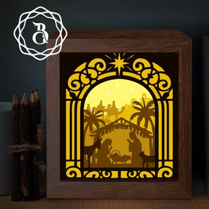 Può includere: Una scatola di luce in legno con un design di carta tagliata gialla e marrone che rappresenta la scena della natività. La scena include una stalla, una mangiatoia, un asino, una mucca, una stella e tre re magi che cavalcano cammelli.