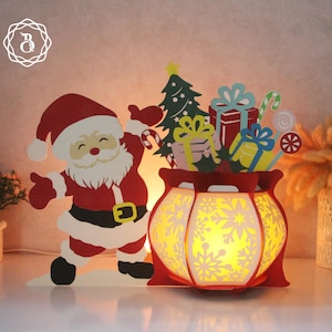 May include: A red and white Santa Claus figure holding a red and white candy cane stands next to a red and white lantern with a snowflake design. The lantern is filled with colorful gifts and candy canes.