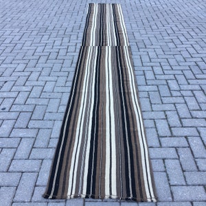 May include: A long, narrow striped rug with brown, black, and white stripes. The rug is made of a woven material and has a fringed edge.
