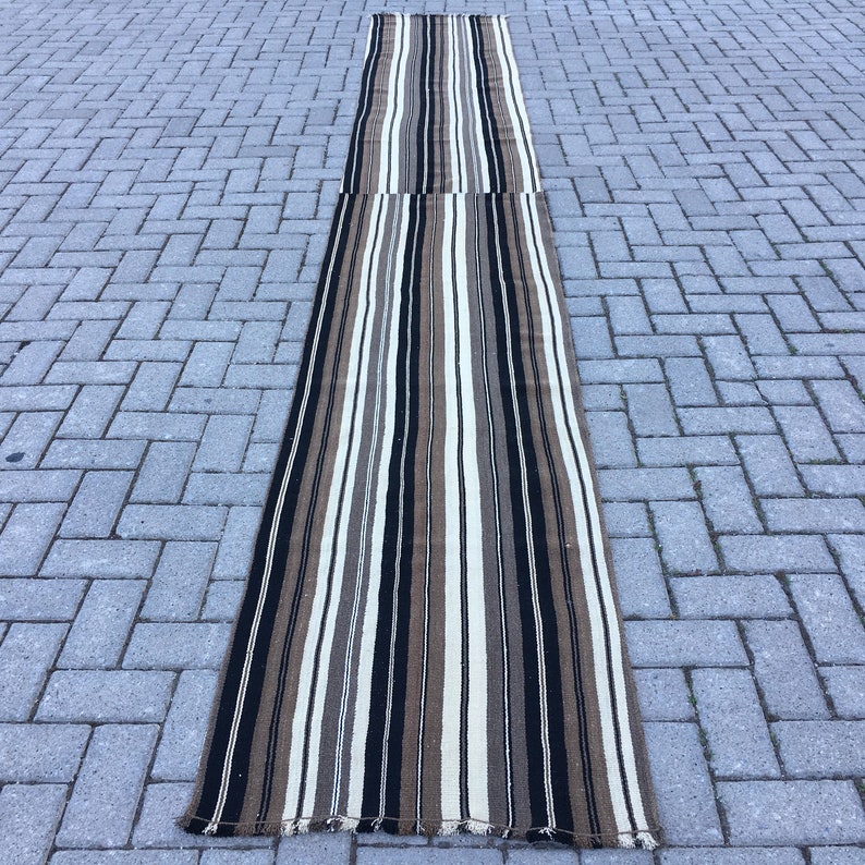 May include: A long, narrow striped rug with brown, black, and white stripes. The rug is made of a woven material and has a fringed edge.