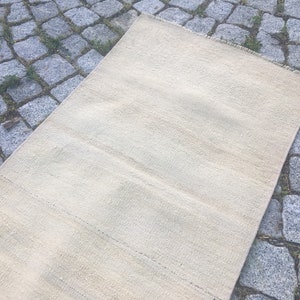 May include: A cream-colored woven rug with a subtle striped pattern. The rug is laid out on a gray stone surface.