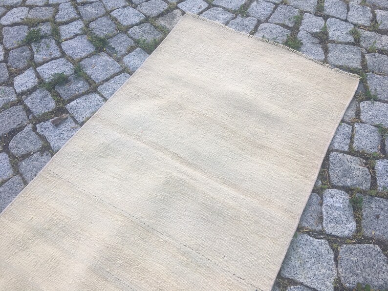 May include: A cream-colored woven rug with a subtle striped pattern. The rug is laid out on a gray stone surface.