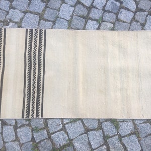 May include: A white woven rug with black geometric patterns along the edges. The rug is laid out on a stone surface.
