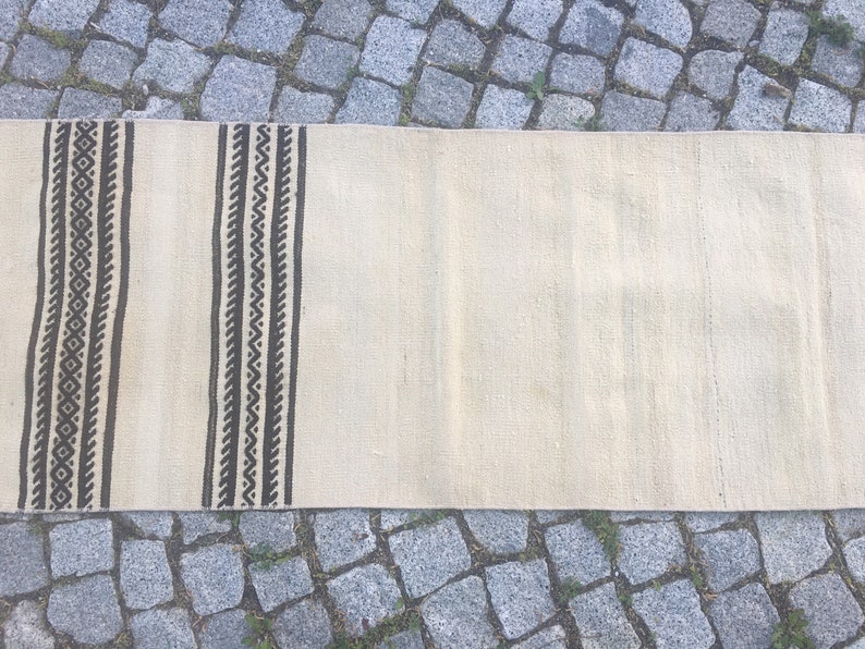 May include: A white woven rug with black geometric patterns along the edges. The rug is laid out on a stone surface.