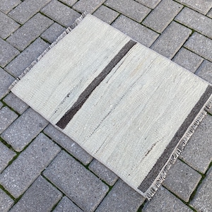 Puede incluir: Una alfombra rayada blanca y gris con un borde marrón. La alfombra tiene un diseño sencillo con una sola raya marrón que recorre el centro.