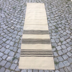 May include: A long, off-white rug with a brown geometric pattern. The rug has a fringe border.