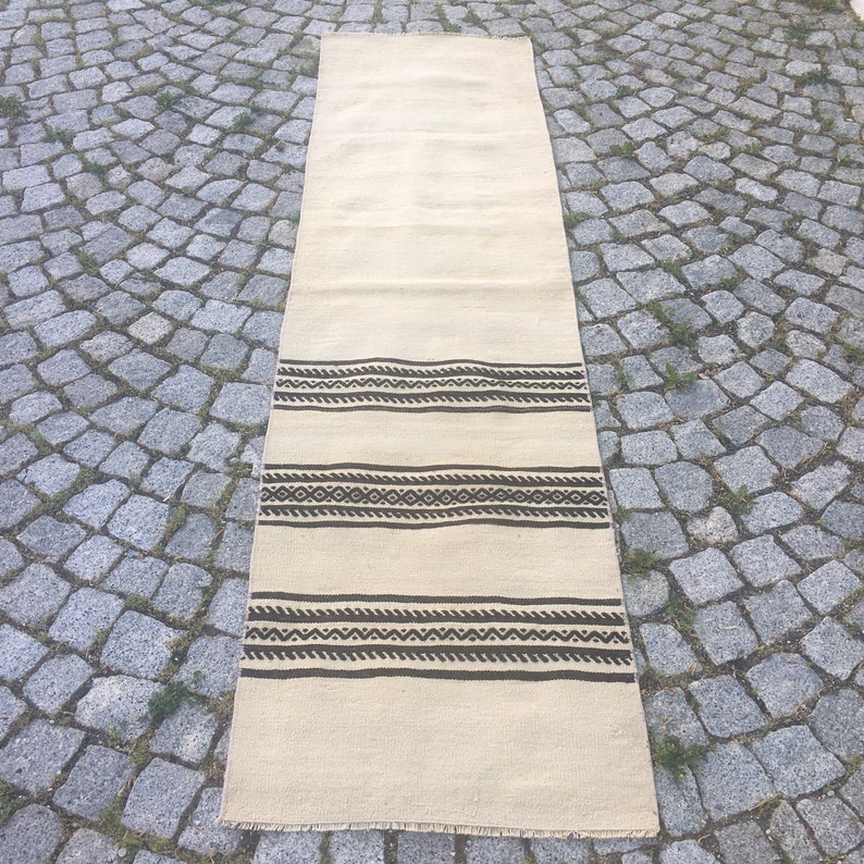May include: A long, off-white rug with a brown geometric pattern. The rug has a fringe border.
