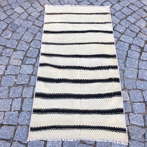 K&ouml;nnte beinhalten: Ein wei&szlig;-schwarz gestreifter Teppich mit Fransenrand. Der Teppich hat ein schlichtes Design mit dicken schwarzen Streifen auf wei&szlig;em Hintergrund.