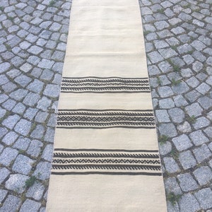 May include: A long, off-white rug with a brown geometric pattern. The rug has a fringe border.