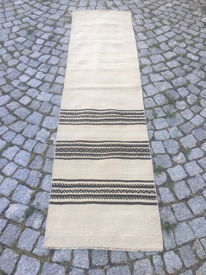 May include: A long, off-white rug with a brown geometric pattern. The rug has a fringe border.
