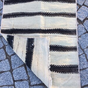 K&ouml;nnte beinhalten: Ein wei&szlig;-schwarz gestreifter Teppich mit Fransenrand. Der Teppich ist in der Mitte gefaltet und zeigt die R&uuml;ckseite.