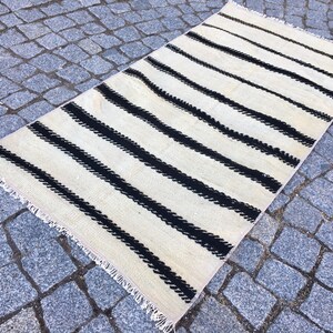 K&ouml;nnte beinhalten: Ein wei&szlig;-schwarz gestreifter Teppich mit Fransenrand. Der Teppich ist aus Wolle und hat eine handgewebte Textur.