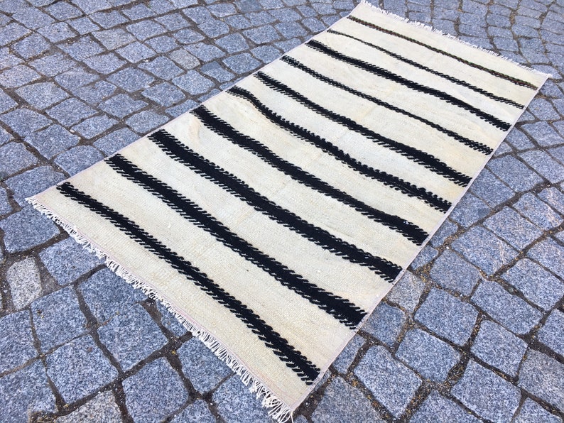 K&ouml;nnte beinhalten: Ein wei&szlig;-schwarz gestreifter Teppich mit Fransenrand. Der Teppich ist aus Wolle und hat eine handgewebte Textur.