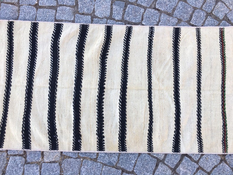 K&ouml;nnte beinhalten: Ein wei&szlig;er und schwarzer gestreifter Teppich mit einem subtilen, geschwungenen Muster in den schwarzen Streifen. Der Teppich hat einen Fransenrand.