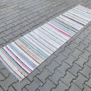 以下が含まれることがあります： 白、グレー、ピンク、茶色など、さまざまな色でストライプ模様が施された、長方形のラグ。 ラグにはフリンジエッジがあり、灰色のレンガの表面に置かれています。 ラグのデザインはシンプルで、ランナーとして使用できます。