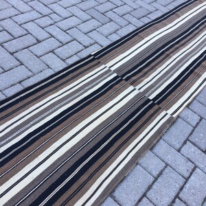 May include: A long, striped rug with black, brown, and white stripes. The rug is made of a woven material and is laid out on a gray brick surface.