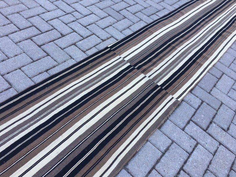 May include: A long, striped rug with black, brown, and white stripes. The rug is made of a woven material and is laid out on a gray brick surface.