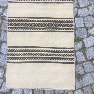 May include: A white rug with black and brown geometric patterns. The rug has a fringe border.