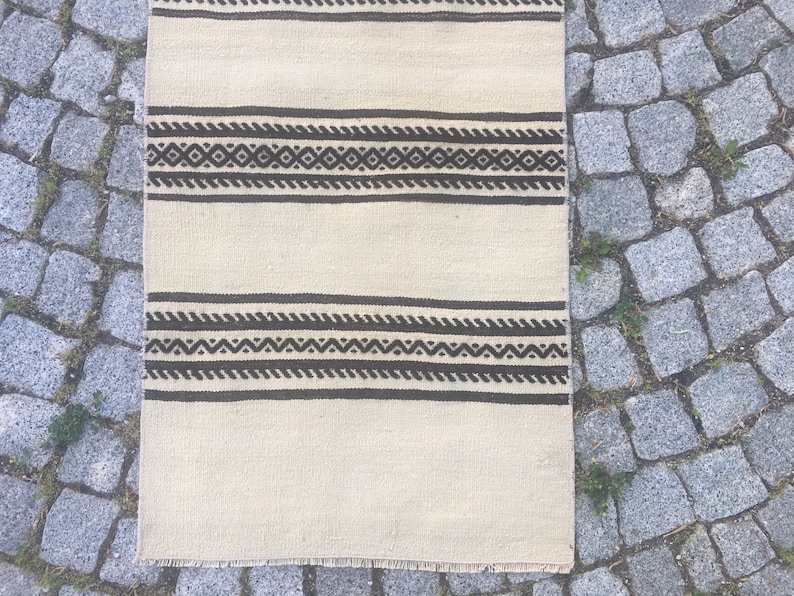 May include: A white rug with black and brown geometric patterns. The rug has a fringe border.