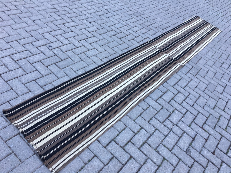 May include: A long, striped rug with black, white, and brown stripes. The rug is made of a woven material and has a fringed edge.