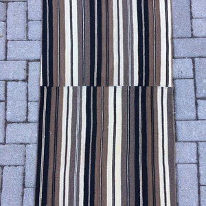 May include: A long, narrow striped rug with alternating bands of black, brown, cream, and white. The rug is woven with a simple, traditional pattern.