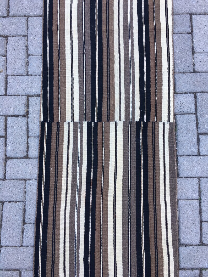 May include: A long, narrow striped rug with alternating bands of black, brown, cream, and white. The rug is woven with a simple, traditional pattern.