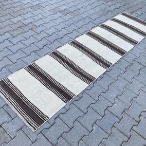May include: A white and brown striped runner rug with a woven texture. The rug has a long, rectangular shape and is laid out on a gray concrete surface.