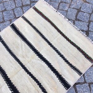 K&ouml;nnte beinhalten: Ein wei&szlig;er Teppich mit schwarzen Streifen und einem gr&uuml;nen und braunen Rand. Der Teppich hat einen Fransenrand.