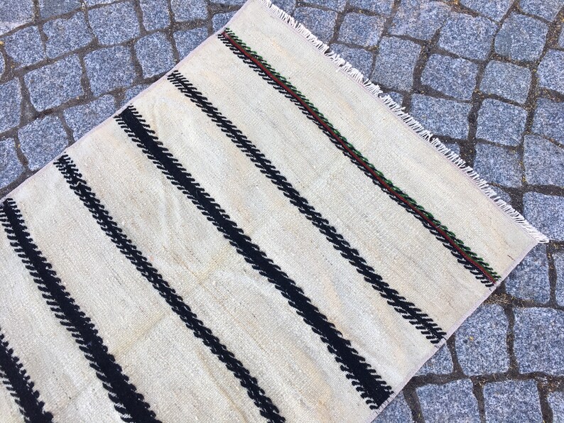K&ouml;nnte beinhalten: Ein wei&szlig;er Teppich mit schwarzen Streifen und einem gr&uuml;nen und braunen Rand. Der Teppich hat einen Fransenrand.