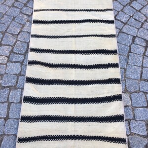 K&ouml;nnte beinhalten: Ein wei&szlig;er und schwarzer gestreifter Teppich mit Fransenrand. Der Teppich hat ein schlichtes Design mit schwarzen Streifen auf wei&szlig;em Hintergrund.