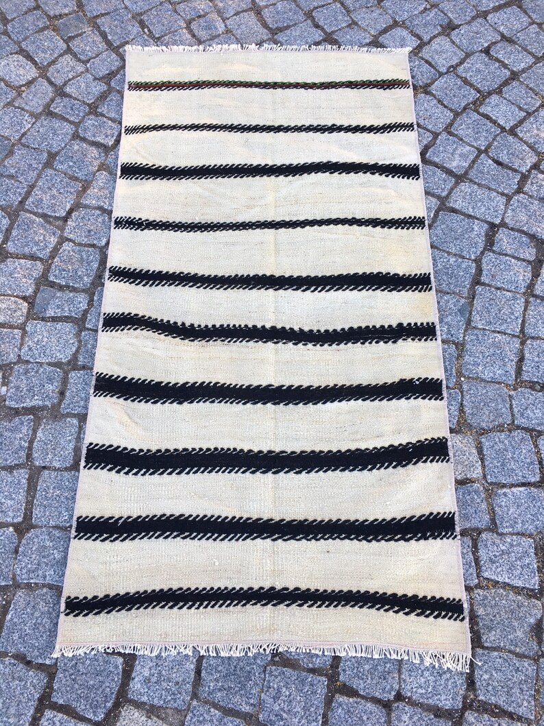 K&ouml;nnte beinhalten: Ein wei&szlig;er und schwarzer gestreifter Teppich mit Fransenrand. Der Teppich hat ein schlichtes Design mit schwarzen Streifen auf wei&szlig;em Hintergrund.