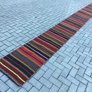 Puede incluir: Una alfombra larga y estrecha con rayas de varios colores, incluyendo rojo, negro, marrón y azul. La alfombra está sobre una superficie de ladrillo gris.