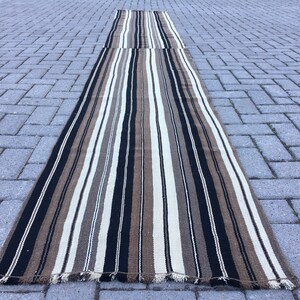 May include: A long, striped rug with black, brown, and white stripes. The rug is made of a woven material and has a fringed edge.