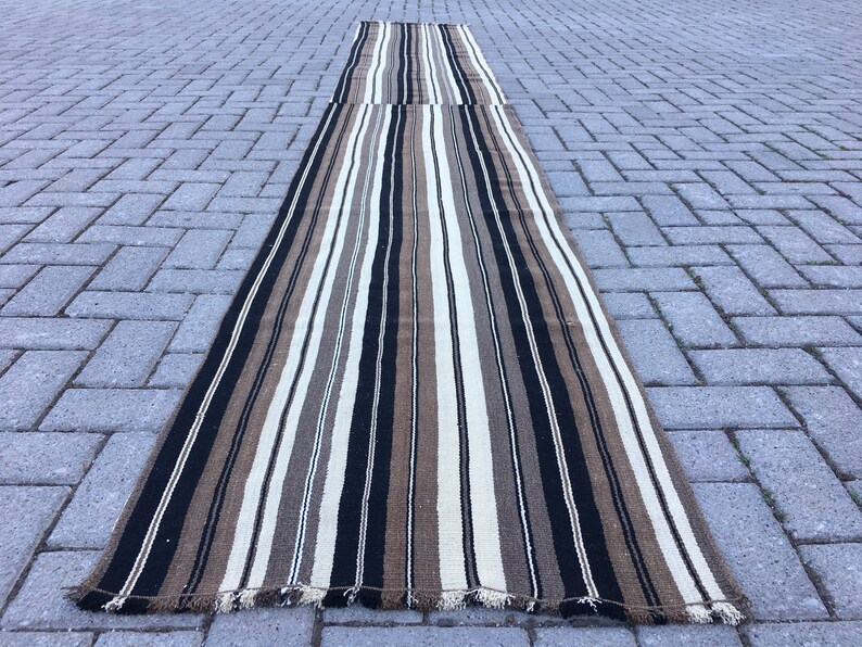 May include: A long, striped rug with black, brown, and white stripes. The rug is made of a woven material and has a fringed edge.