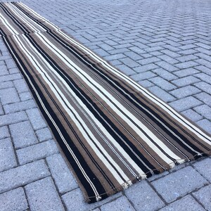 May include: A long, striped rug with black, white, and brown stripes. The rug is made of a woven material and has a fringed edge.