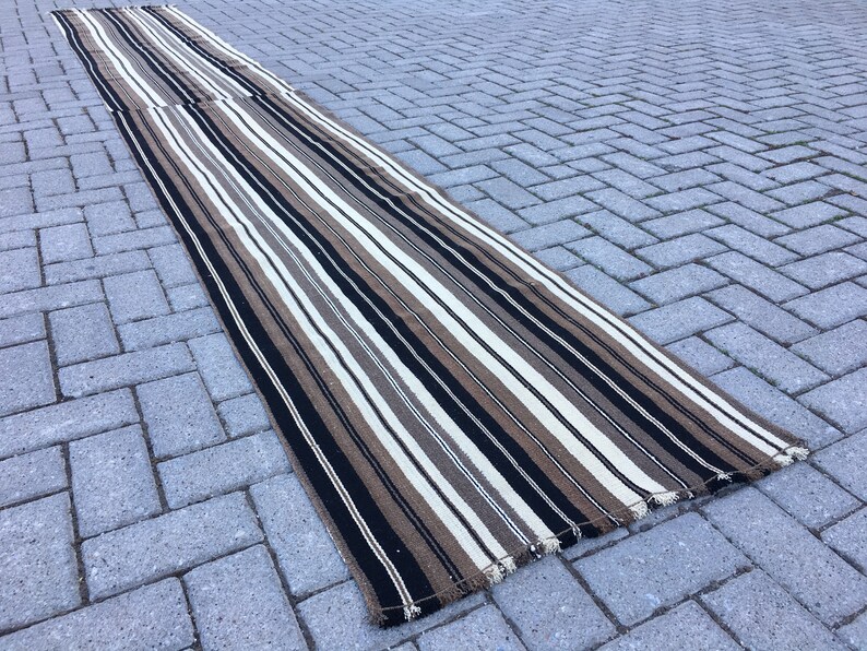 May include: A long, striped rug with black, white, and brown stripes. The rug is made of a woven material and has a fringed edge.